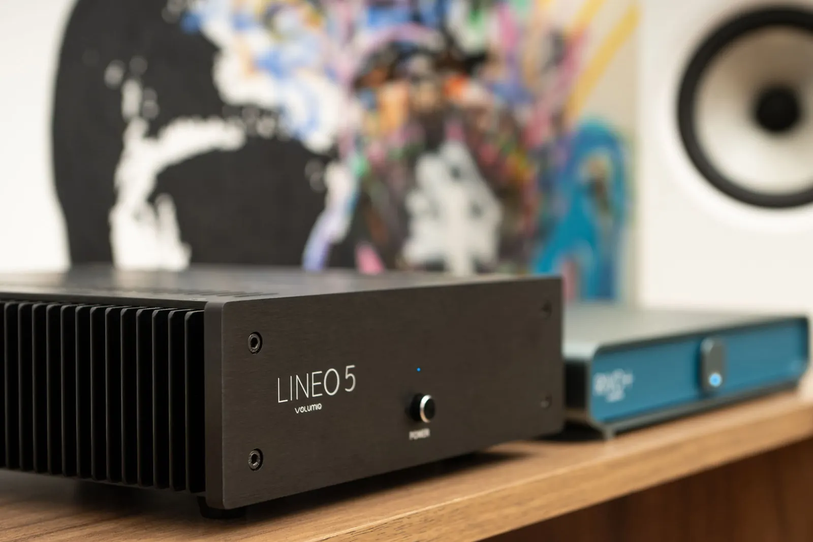 Lineo5 on a wooden shelf, with heatsink fins along its side, next to a Volumio device with a blue front. A colorful painting and a white speaker are behind them.