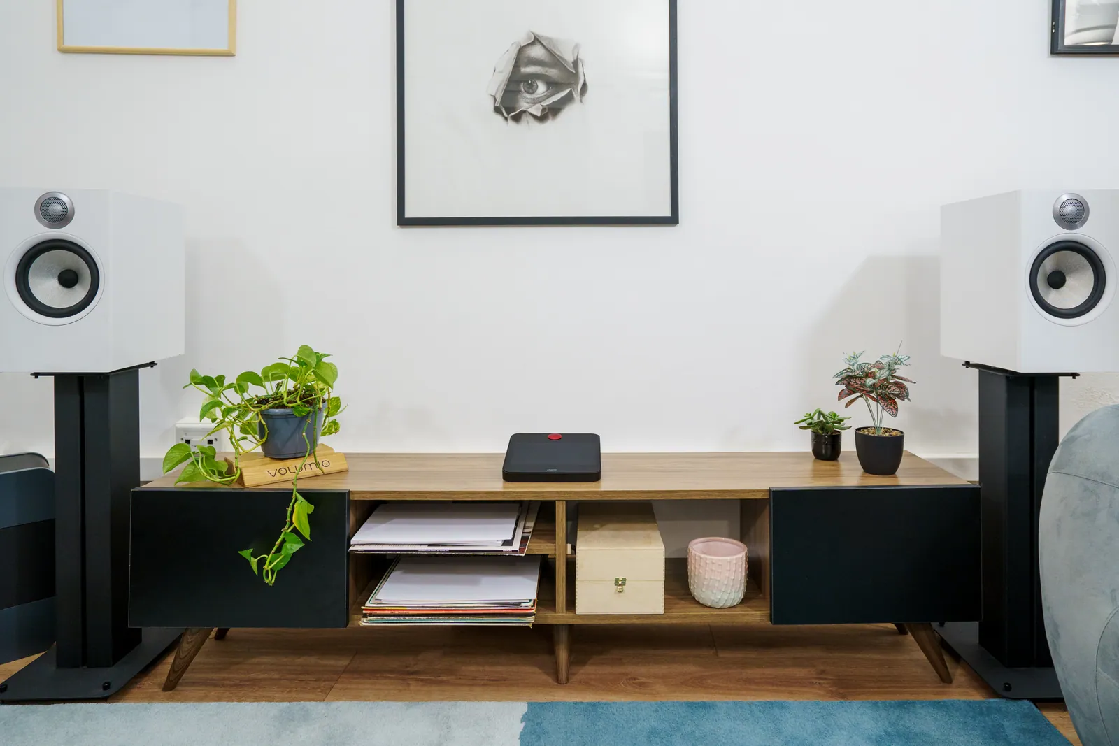 Motivo on a wooden sideboard between two white bookshelf speakers on stands, in a living room with plants.