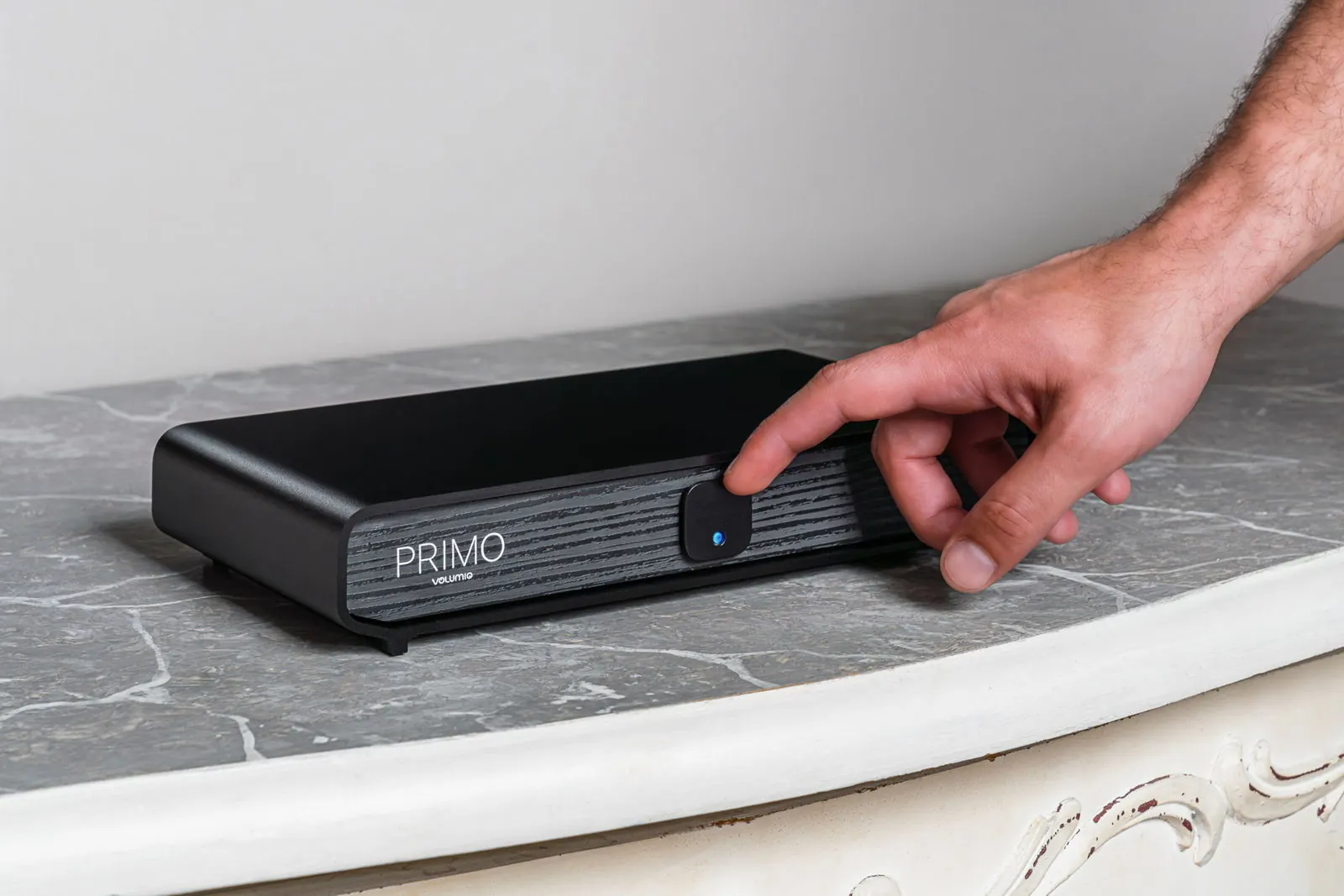 A hand reaching for the button on the front of Primo V3, which sits on a grey marble top.