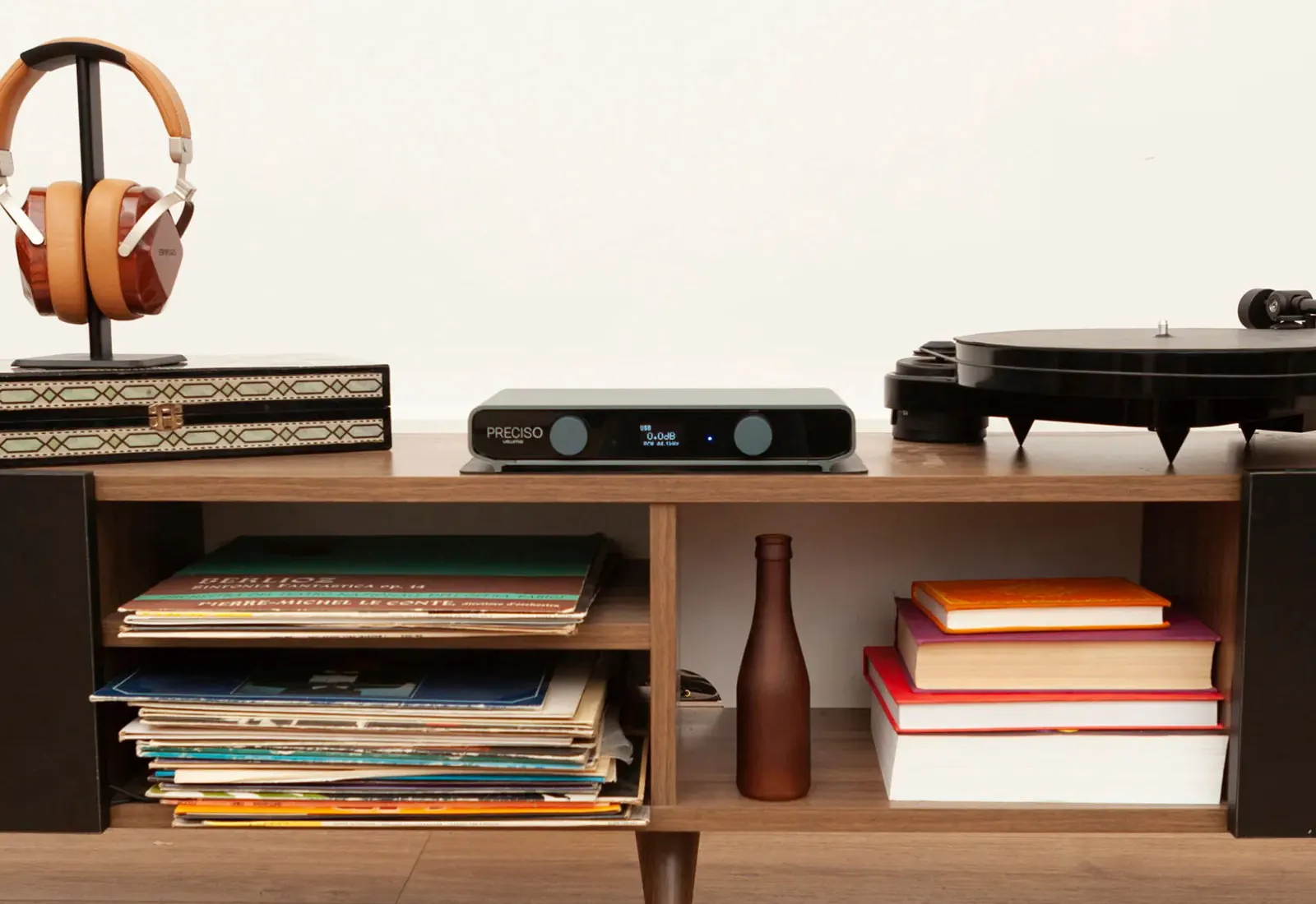 Preciso on a wooden sideboard between headphones on a stand and a turntable, with records, books and a brown bottle on the shelves below.