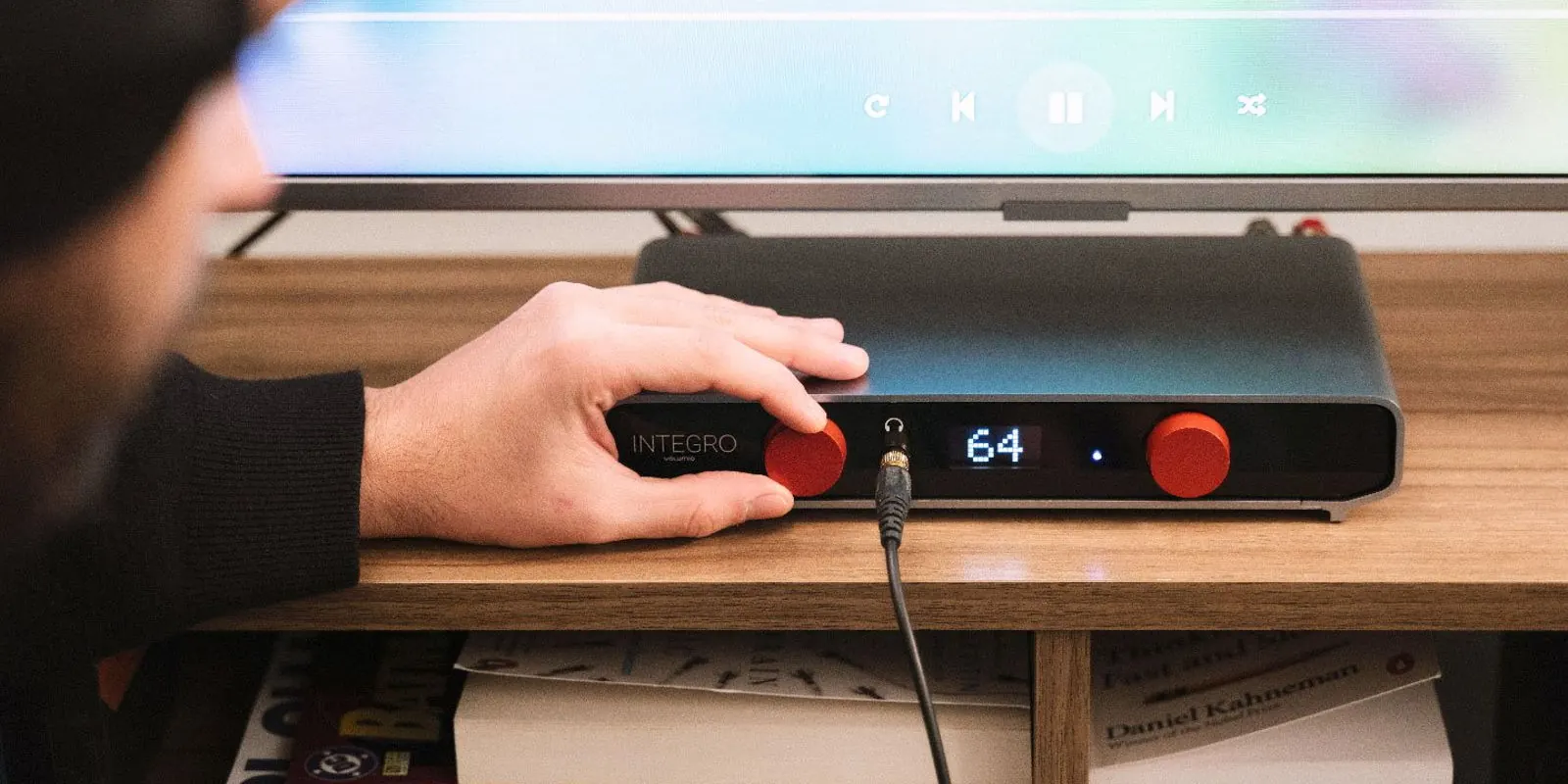 A listener turns a knob on Integro, with a headphone cable plugged into the front and a TV showing playback controls behind it.