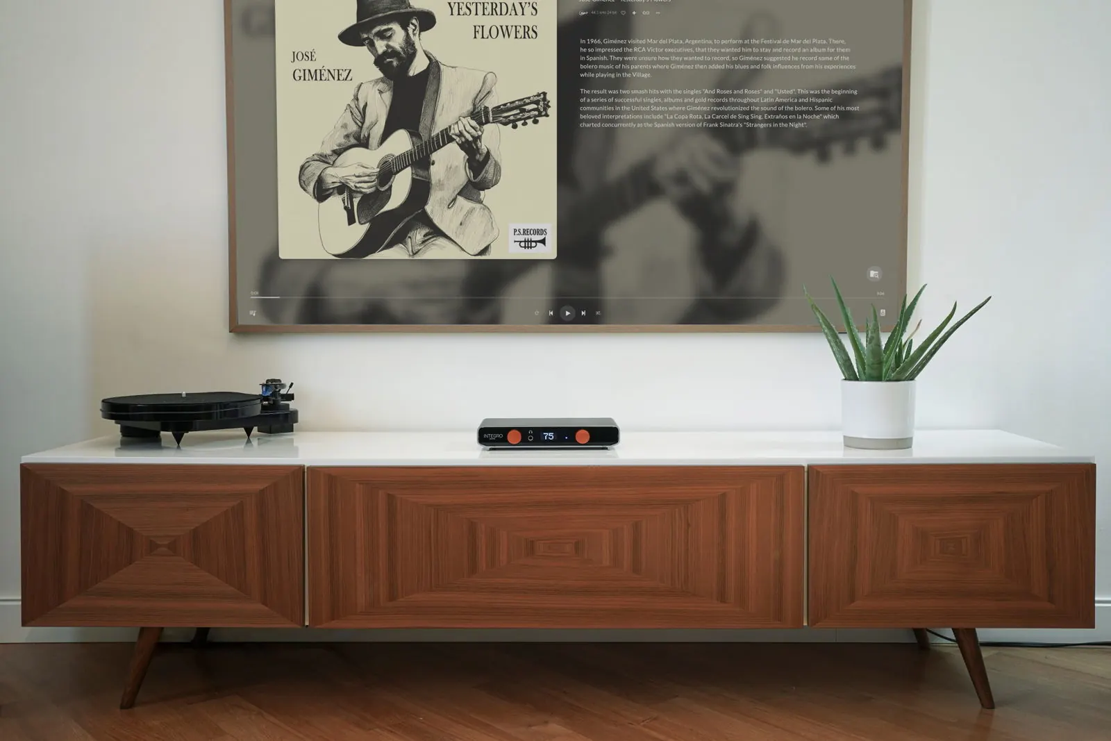 A wooden sideboard with a turntable, Integro and a plant, under a wall TV that shows album art, an artist biography and playback controls.