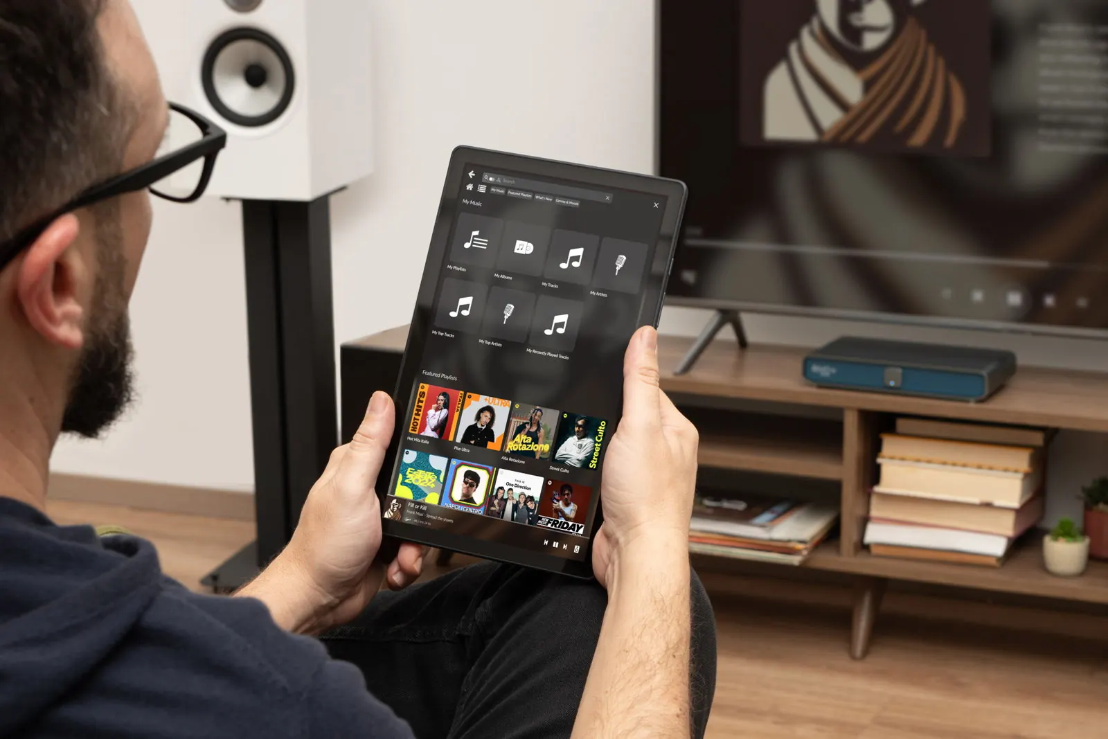 A listener holding a tablet that shows a music library, with Rivo Plus on a sideboard under the TV behind him.